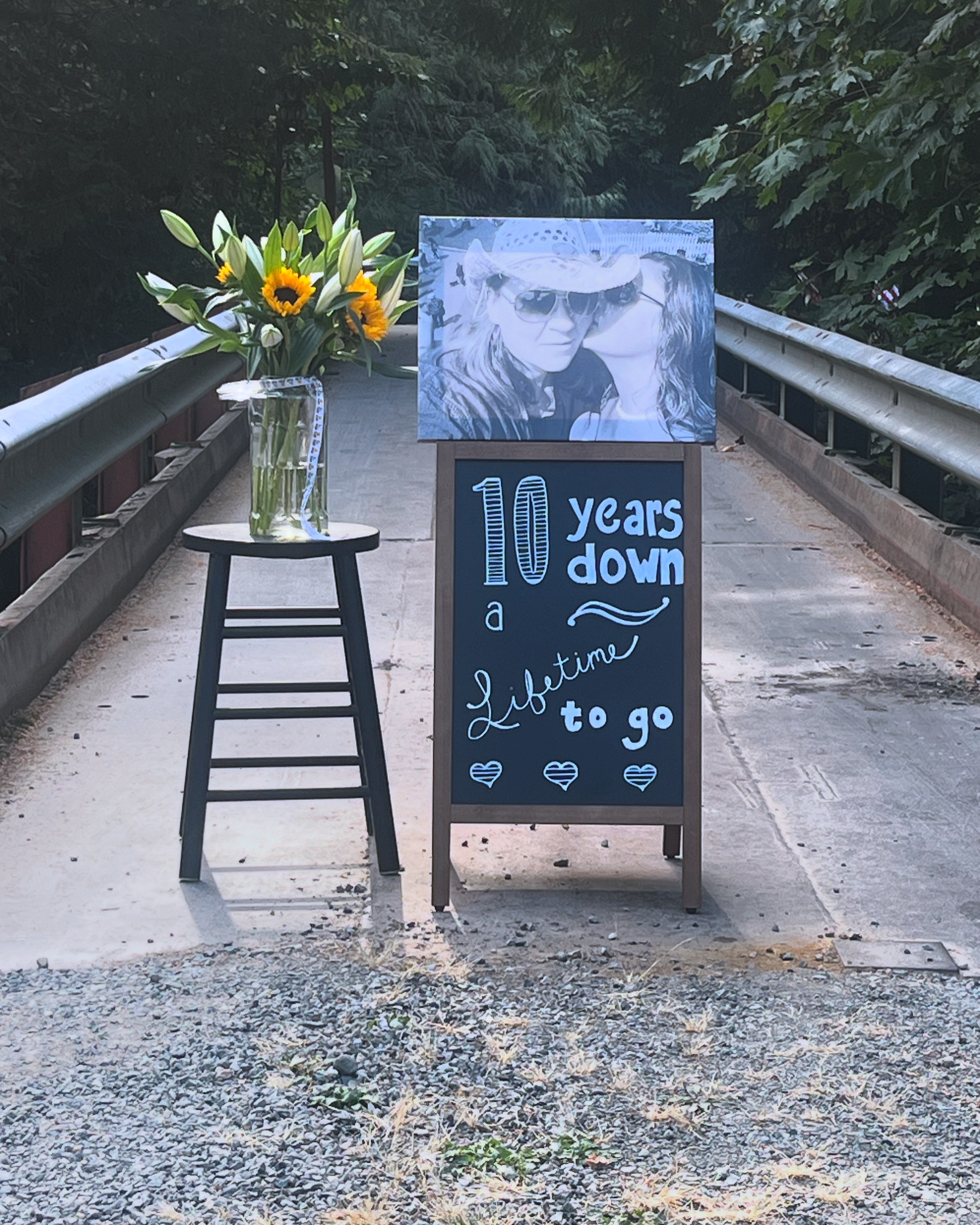Outdoor anniversary display on a bridge with chalkboard sign reading '10 years down a lifetime to go', a portrait photo, and sunflowers in a vase on a stool